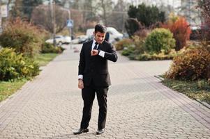 elegante hombre de negocios indio del sur de Asia con traje negro mirando sus relojes inteligentes a mano. foto