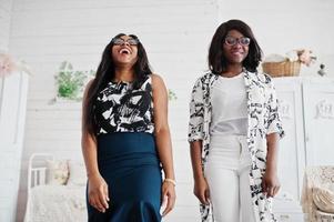 Two african woman friends wear on eyeglasses posed indoor white room. Laughing and having fun together. photo