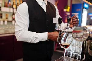 Close up hands of african american bartender at bar pouring from tap fresh beer into the glass in pub. photo