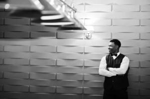 joven apuesto hombre africano vestido con camisa blanca, chaleco negro y corbata de moño posado contra la pared violeta púrpura. foto