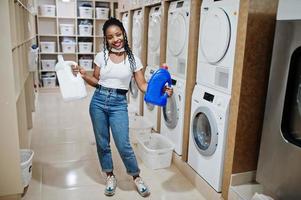 alegre mujer afroamericana sostiene detergente cerca de la lavadora en la lavandería de autoservicio. foto