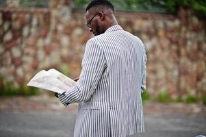 hombre afroamericano rico y elegante con chaqueta y pantalones blancos, anteojos lee la revista. foto