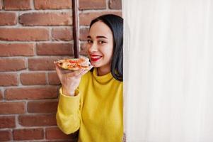 chica morena divertida en suéter amarillo comiendo pizza en el restaurante. foto