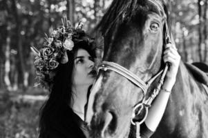 Mystical girl in wreath wear in black with horse in wood. photo