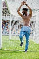 Handsome sexy african american muscular bare torso man at jeans overalls posed at green grass agasinst football gates of stadium field. Fashionable black man portrait. photo