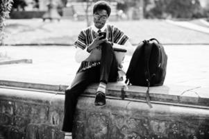 African man student in traditional clothes and glasses with backpack, working with laptop and mobile phone outdoor. photo