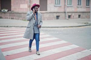 Stylish African American man model in gray coat, jacket tie and red hat walking on crosswalk and speaking on mobile phone. photo