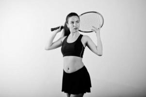 Black and white portrait of beautiful young woman player in sports clothes holding tennis racket while standing against white background. photo