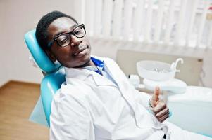 African american male doctor in glasses having rest after hard working day at dentist chair in dental clinic. He is satisfied of result and show thumb up. photo