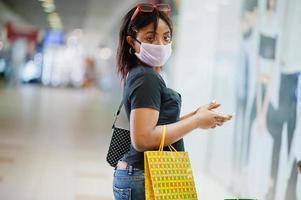 African american woman wearing face protective medical mask for protection from virus disease with mobile phone in mall during coronavirus pandemia. photo