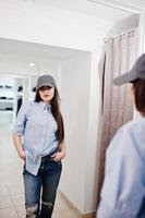 hermosa chica morena en la boutique de la tienda de ropa en ropa casual y gorra contra el espejo. foto