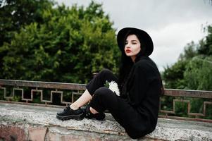 chica sensual todo en negro, labios rojos y sombrero. mujer gótica dramática sostiene una flor de crisantemo blanco. foto