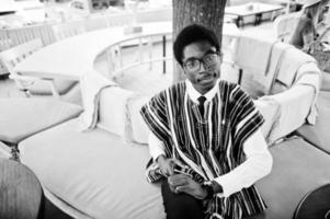 African man in traditional clothes and glasses sitting at outdoor caffe with cell phone. photo