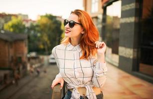 atractiva mujer pelirroja con gafas de sol, vestida con una blusa blanca posando en la calle contra un edificio moderno. foto