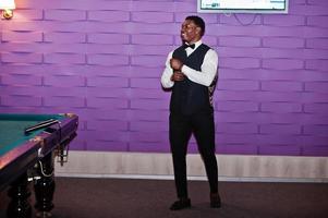 Young handsome african man wearing white shirt, black vest and bow tie standing against pool billiard tables. photo