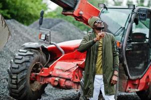 Stylish african american man in hat and sunglasses posed outdoor in rain against tractor with a bucket. photo