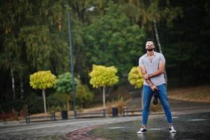 hombre de barba árabe alto de moda usa camisa, jeans y gafas de sol con paraguas posado bajo la lluvia en la plaza del parque. foto