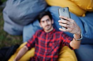 joven estudiante indio con camisa a cuadros y jeans sentado y relajado en almohadas al aire libre y haciendo selfie. foto