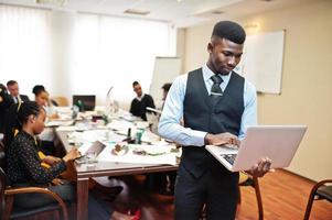 Face of handsome african business man, holding laptop on the background of business peoples multiracial team meeting, sitting in office table. photo