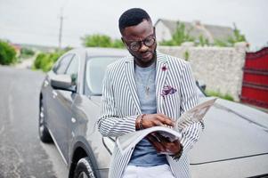 hombre afroamericano rico y elegante en blazer y pantalones blancos, anteojos lee la revista contra su auto todoterreno. foto