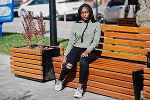 City portrait of positive young dark skinned female wearing green hoody sitting on bench. photo