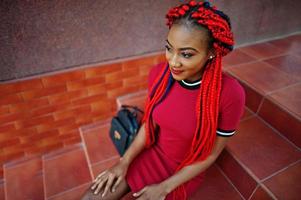 Cute and slim african american girl in red dress with dreadlocks and backpack posed outdoor, sitting on stairs. Stylish black model. photo