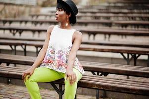 Amazing african american model woman in green pants and black hat posed at bench. photo