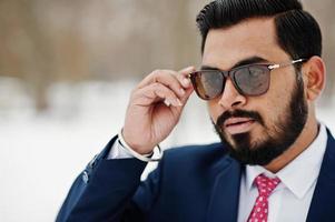 primer plano retrato de elegante hombre de negocios de barba india en traje y gafas de sol posado en el día de invierno al aire libre. foto