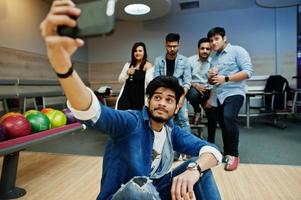 Group of five south asian peoples having rest and fun at bowling club. Making selfie by phone and holding cold soda drinks from glass bottles. photo