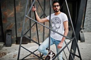 Street style arab man in eyeglasses with longboard posed inside metal pyramid construction. photo