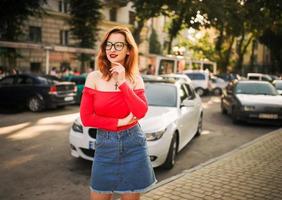 atractiva mujer pelirroja con anteojos, blusa roja y falda de jeans posando en la calle contra un auto deportivo blanco. foto