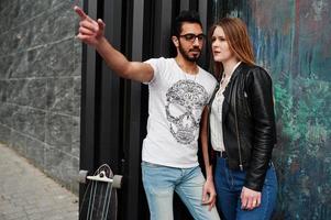 Cool pareja multirracial posando contra la pared con longboard. El novio árabe le muestra algo a su chica europea. foto
