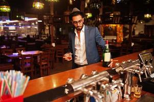 Handsome well-dressed arabian man with glass of whiskey and cigar posed at pub. photo