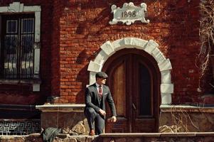 Portrait of retro 1920s english arabian business man wearing dark suit, tie and flat cap standing against old brick house 1898 year. photo