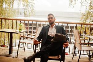 Handsome well-dressed arabian man smoke cigar with glass of whiskey at balcony of pub. photo