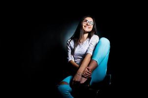 Portrait of an attractive young woman in white top and blue pants sitting posing with her glasses in the dark. photo