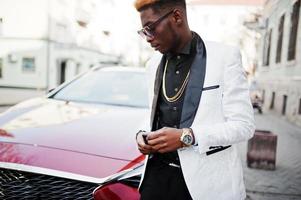 Chic handsome african american man in white suit against red luxury car with mobile phone at hand. photo