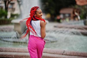 chica afroamericana de moda en pantalones rosas y rastas rojas posadas al aire libre contra fuentes. foto