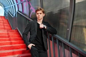 Young stylish macho boy in black jacket posed outdoor of street. Amazing model man at red stairs tonnel. photo