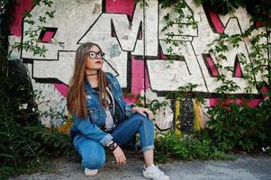 chica hipster casual con estilo en ropa de jeans y gafas contra una gran pared de graffiti. foto