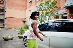 Increíble mujer modelo afroamericana con pantalones verdes y sombrero negro posada cerca de la puerta blanca del auto. foto