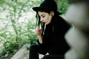 chica fumadora sensual todo en negro, labios rojos y sombrero. mujer dramática gótica fumando un cigarrillo delgado. foto