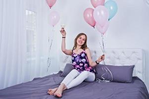Happy girl with colored balloons on bed at room with glasses of champagne. Celebrating birthday theme. photo