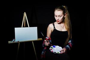 Beautiful woman artist painter with brushes and oil canvas posing in studio isolated on black. photo