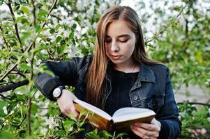 jovencita morena en jeans contra el árbol de flor de primavera lee el libro. foto