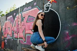 Back of buttocks stylish casual hipster girl in cap, sunglasses and jeans wear, listening music from headphones of mobile phone against large graffiti wall with large tnt bomb. photo