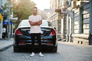 Stylish indian beard man at sunglasses and pink t-shirt against luxury car. India rich model posed outdoor at streets of city. photo