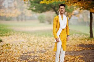 Indian stylish man in yellow traditional clothes with white scarf posed outdoor against autumn leaves tree. photo