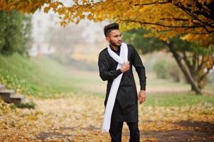 Indian stylish man in black traditional clothes with white scarf posed outdoor against yellow autumn leaves tree. photo