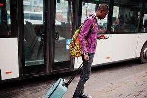 African american man in checkered shirt, with suitcase and backpack. Black man traveler against bus. photo